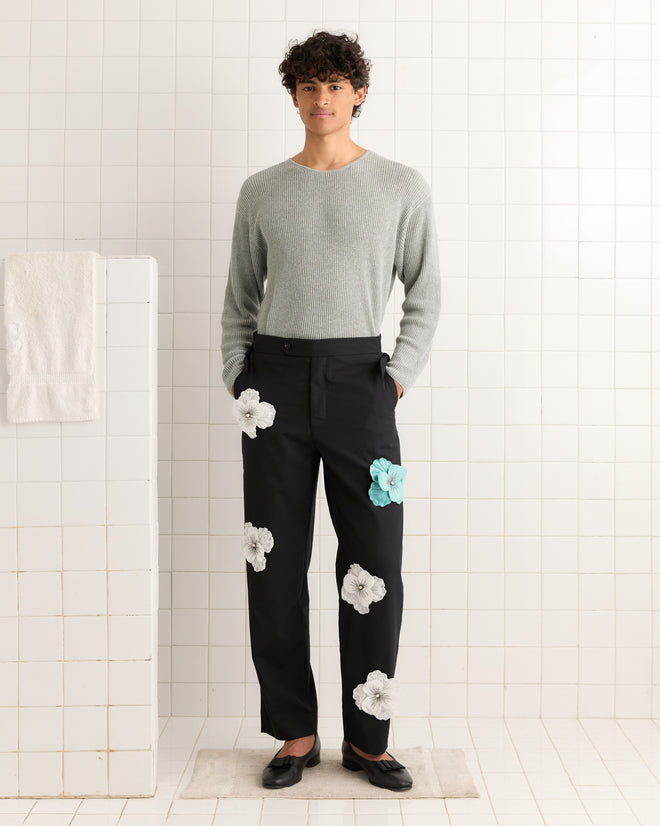 A person in a gray sweater and Tropical Wool Pansy Trousers with large antique flower appliqué stands in a tiled room.