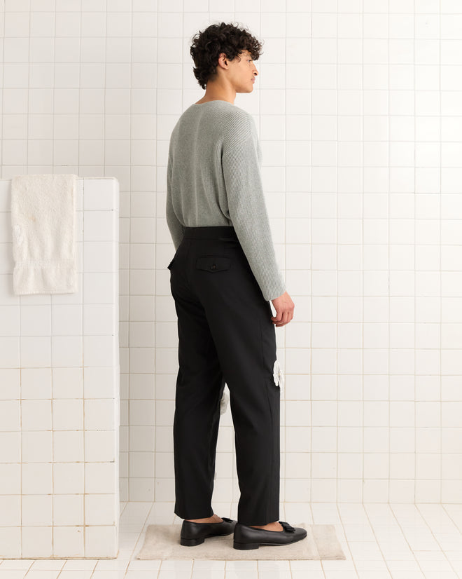 A person in a gray 100% wool sweater and black Tropical Wool Pansy Trousers stands sideways in a white-tiled room.