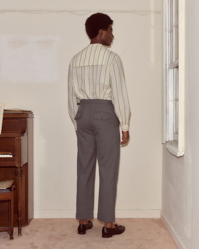 A man in a striped shirt and Tropical Wool Side-Buckle Trousers - Grey stands facing a corner in a sparsely furnished room.