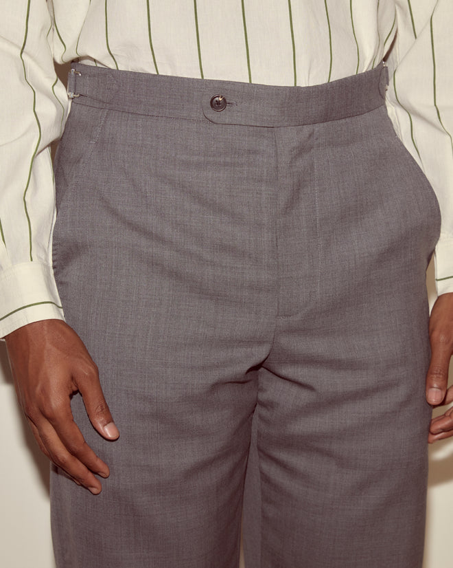Model wears Tropical Wool Side-Buckle Trousers in grey, paired with a cream shirt featuring green stripes, shown from waist to mid-thigh.