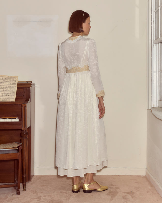 A woman in the cream Trousseau Lace Dress stands by a piano, facing away, in a softly lit room.