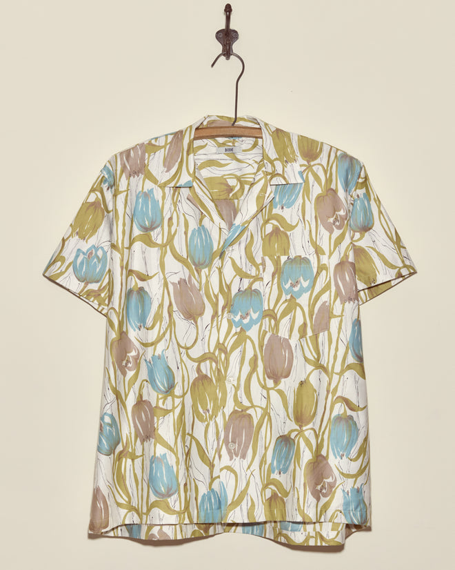 The Tulip Watercolor Shirt - M/L is a unique short-sleeved shirt featuring blue, brown, and green tulip patterns, crafted in New York and displayed on a metal hook.