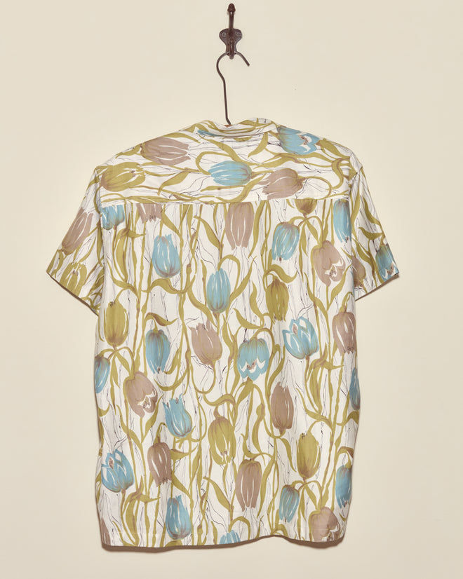 The Tulip Watercolor Shirt - M/L, a unique short-sleeve deadstock top with blue, green, and beige tulip motifs, hangs on a hook against a cream-colored wall.