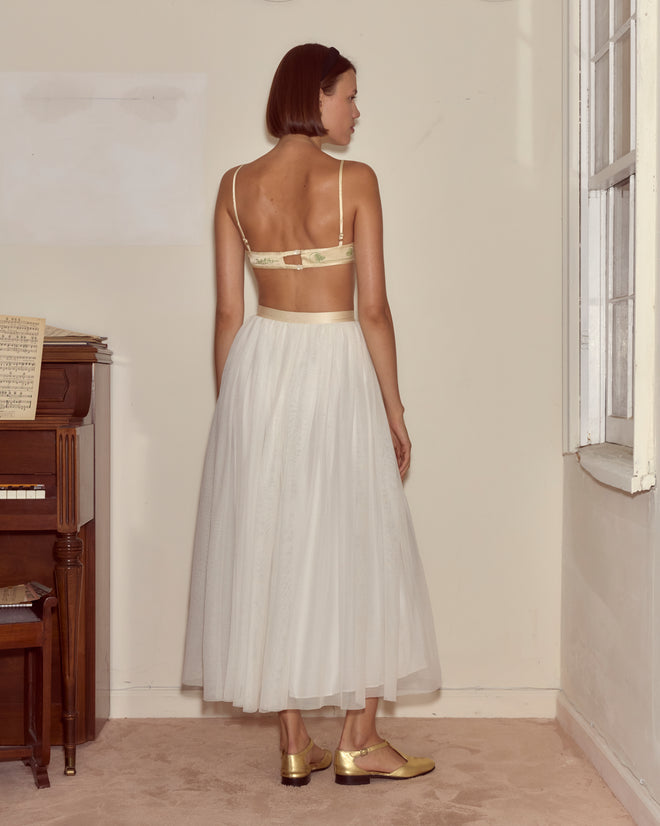 A woman wearing the Tulle Tea Skirt and gold shoes stands facing away in a lightly furnished room near a window.