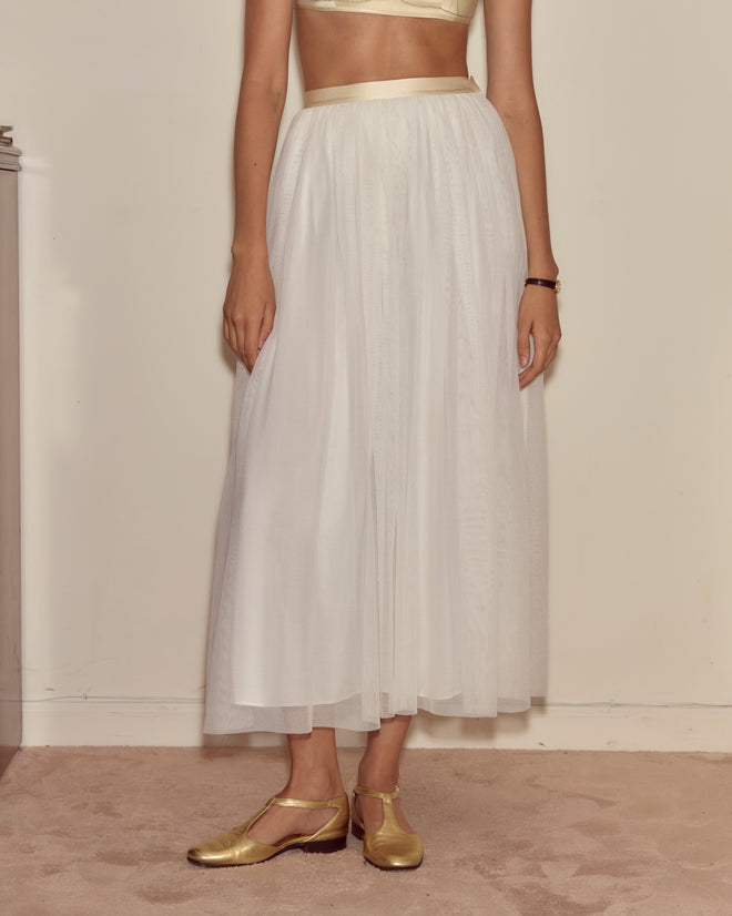 A person wears the Tulle Tea Skirt, a vintage-inspired white tulle skirt, paired with a light top and gold shoes, standing on a beige carpet before a cream wall.