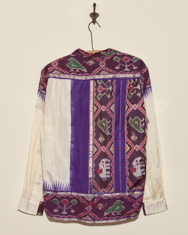 The Turaco Silk Shirt - L/XL, featuring unique purple, white, and multicolor designs, hangs on a hook against the wall.