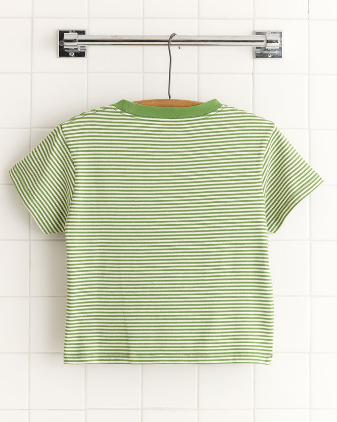 The Truro Stripe Cropped Tee, inspired by 1950s vintage fashion, features green and white stripes with a green collar and is made from soft cotton. It hangs elegantly on a hanger against a tiled wall.