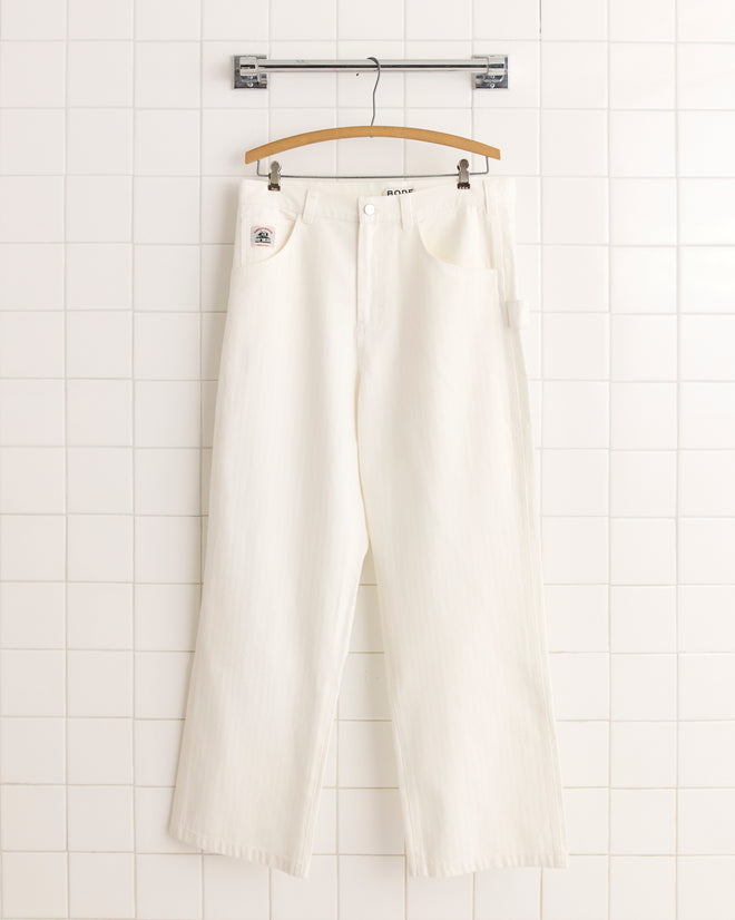 The Twill Knolly Brook Trousers in white, featuring a vintage workwear silhouette, hang from a wooden hanger against a white tiled wall.