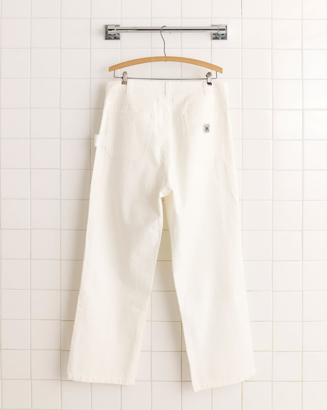 The Twill Knolly Brook Trousers - White, made from 100% cotton, hang against a tiled white wall. These workwear pants feature a vintage silhouette and are displayed from the back for a classic look.