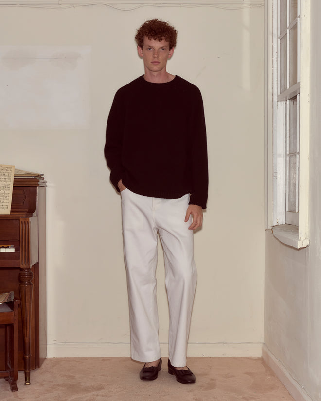 A man stands indoors by a piano wearing a black sweater, the Twill Knolly Brook Trousers in white with a hammer loop, and black shoes.