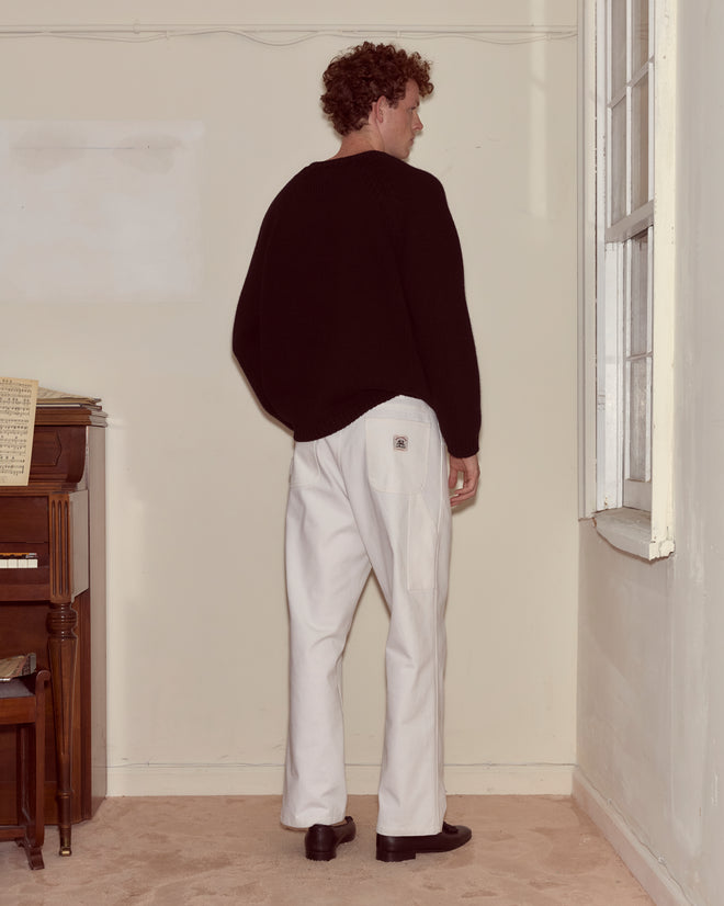 A man stands indoors facing a wall, wearing a black sweater, black shoes, and white Twill Knolly Brook Trousers with a hammer loop.
