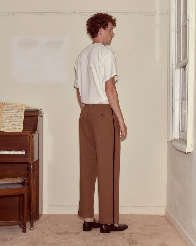 A man in a white shirt and Twill Track Pants - Brown stands sideways near a piano, facing a window in a beige room.