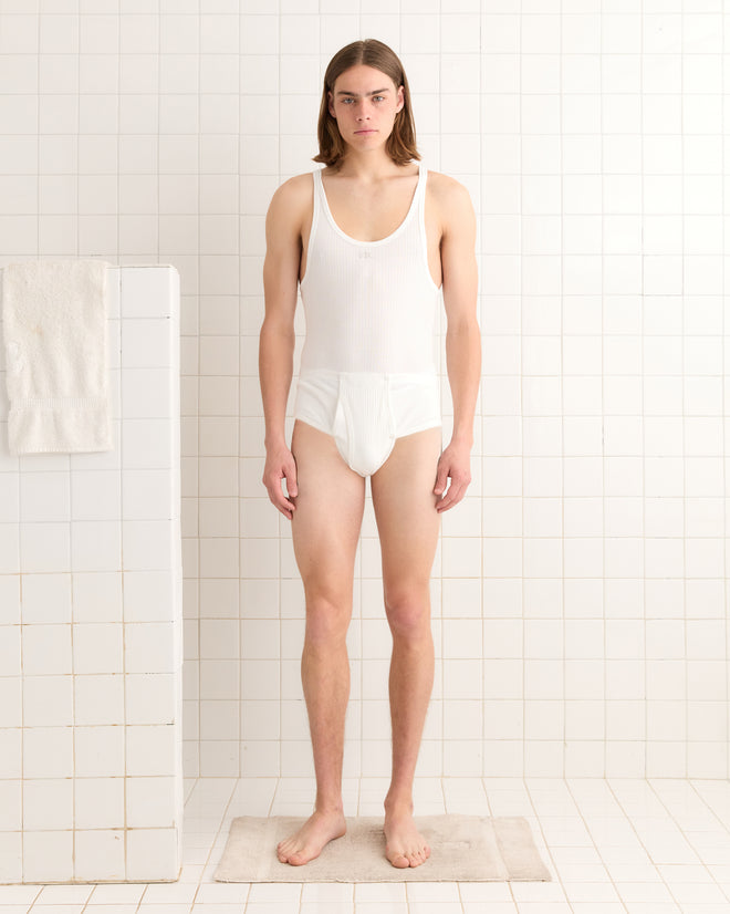 A person with long hair stands barefoot in a white Undersuit, reminiscent of historical reproduction attire, on a bath mat in a tiled bathroom.
