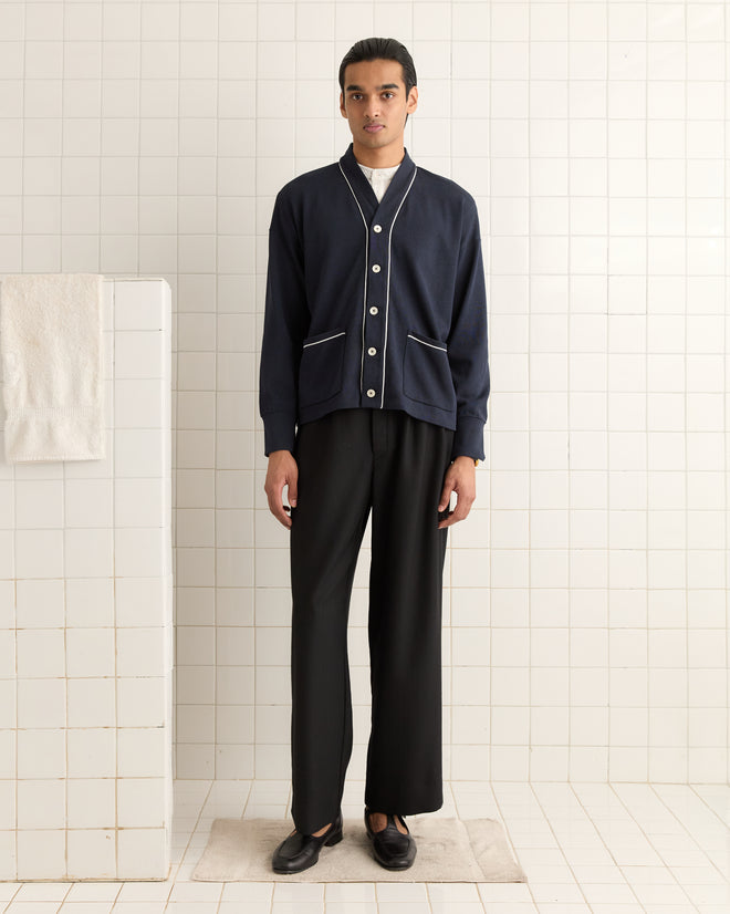 A person wearing a Uniform Cardigan with black pants is standing on a beige mat in a tiled room.