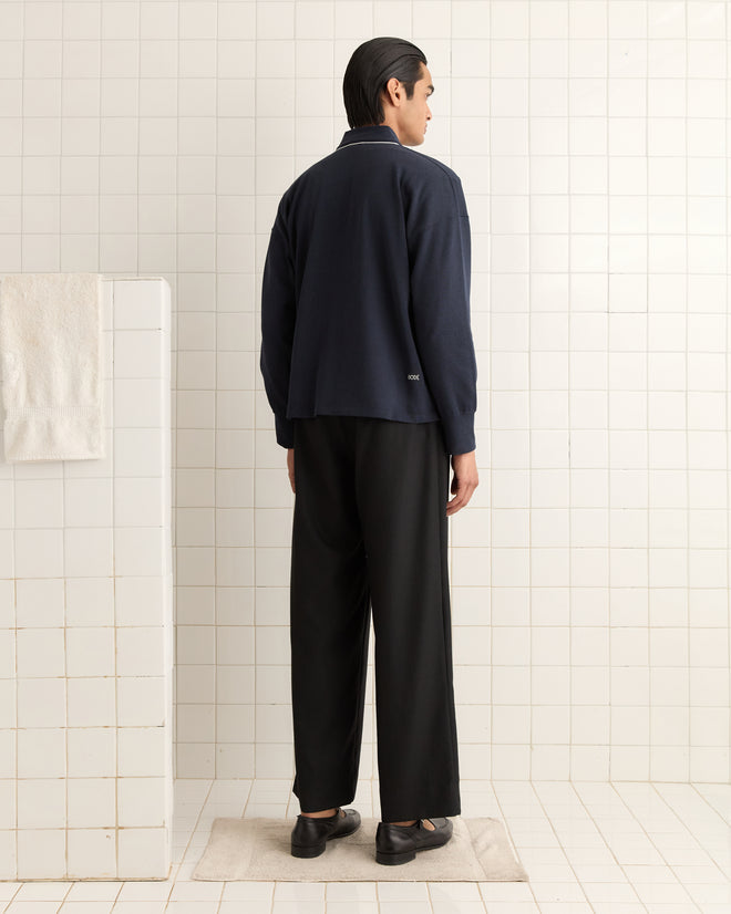 A person wearing a navy shirt and black pants stands on a beige mat in a tiled bathroom, emphasizing the heavyweight fabric of the Uniform Cardigan draped over their shoulders.
