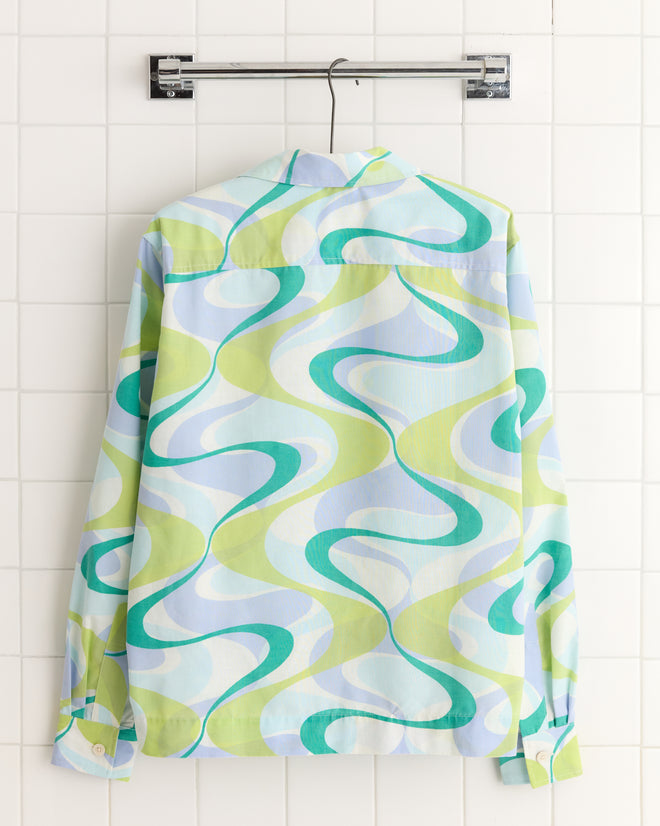 The Upstream Creek Shirt, a long-sleeve style with a green and blue abstract pattern, hangs on a hanger against white tiles. It's crafted from unique deadstock yardage.