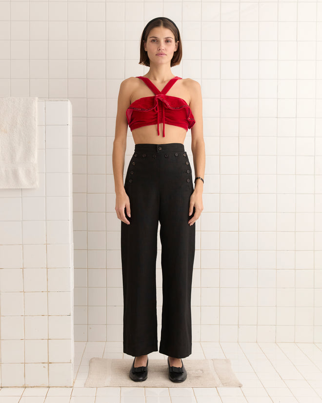 In a tiled room, a person stands wearing the Velvet Ruffle Bra, inspired by 1950s red velvet fashion with adjustable straps, paired with black pants, their hands resting calmly by their sides.