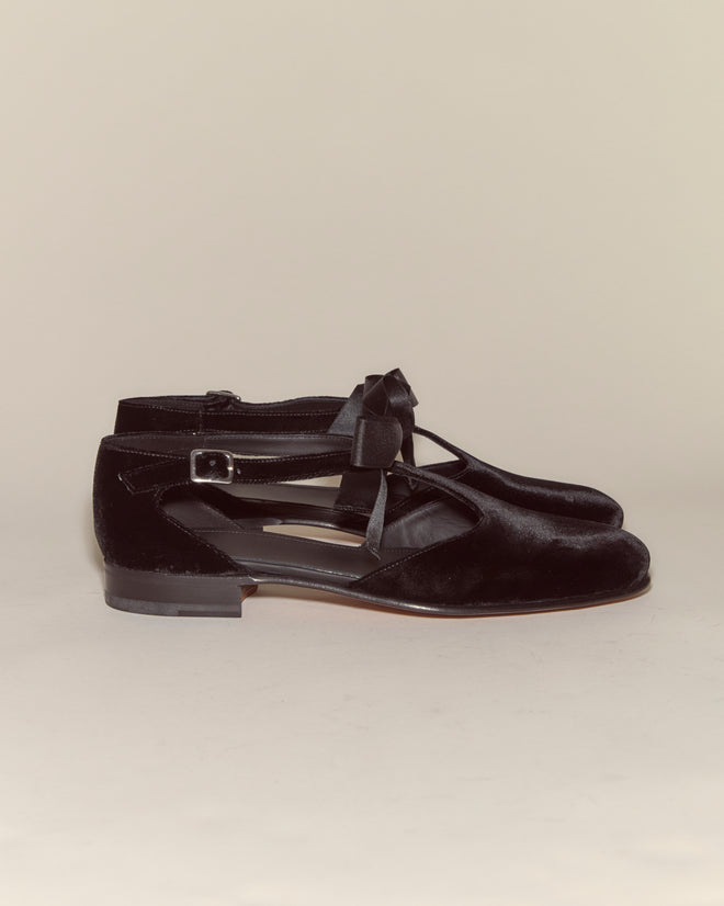 The Velvet Theater Shoes - Black are black velvet Mary Jane flats with a bow, buckle strap, and subtle T-strap detail, evoking midcentury dancing shoe charm against a neutral background.