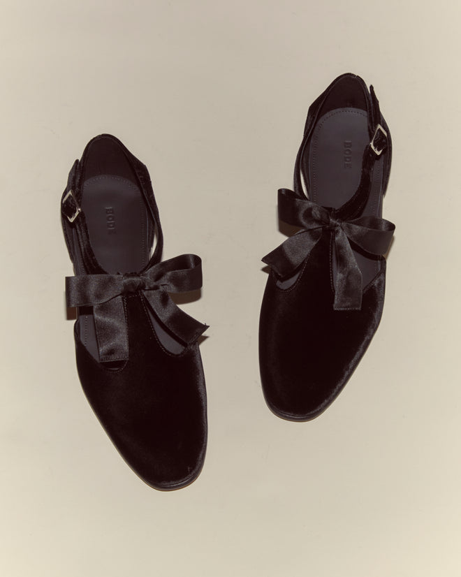A top view of the Velvet Theater Shoes - Black, featuring elegant ribbon bows and a charming T-strap detail, set against a light background.