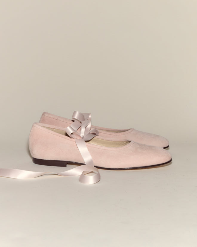 A pair of Musette Flats - Pink, featuring pale pink suede and long satin ribbon laces, rests on a plain background.
