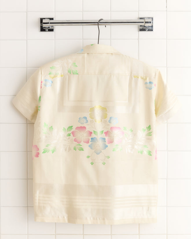 The Waterlily Stencil Shirt - XS, a short-sleeve shirt with cream and soft pastel floral patterns, hangs on a metal hanger against a tiled wall.