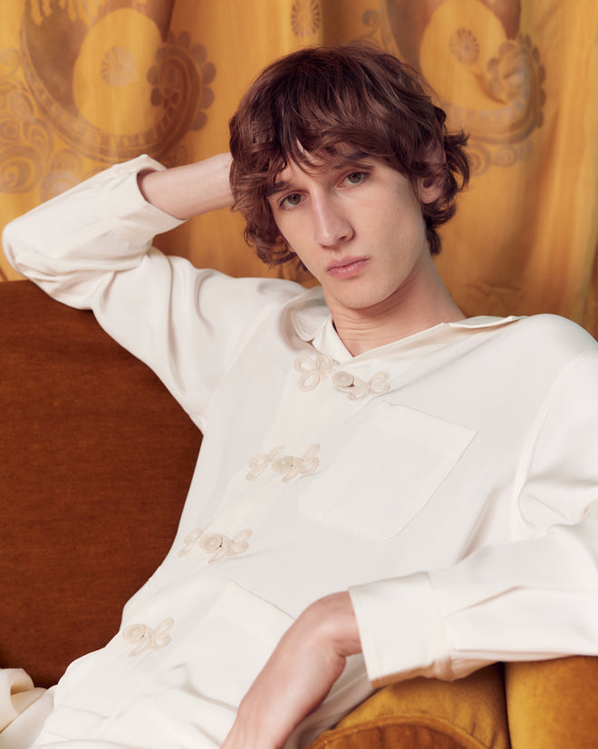 A young person with wavy brown hair sits on a gold couch, posing with one hand behind their head while wearing the Trillium Long Sleeve Shirt in White.