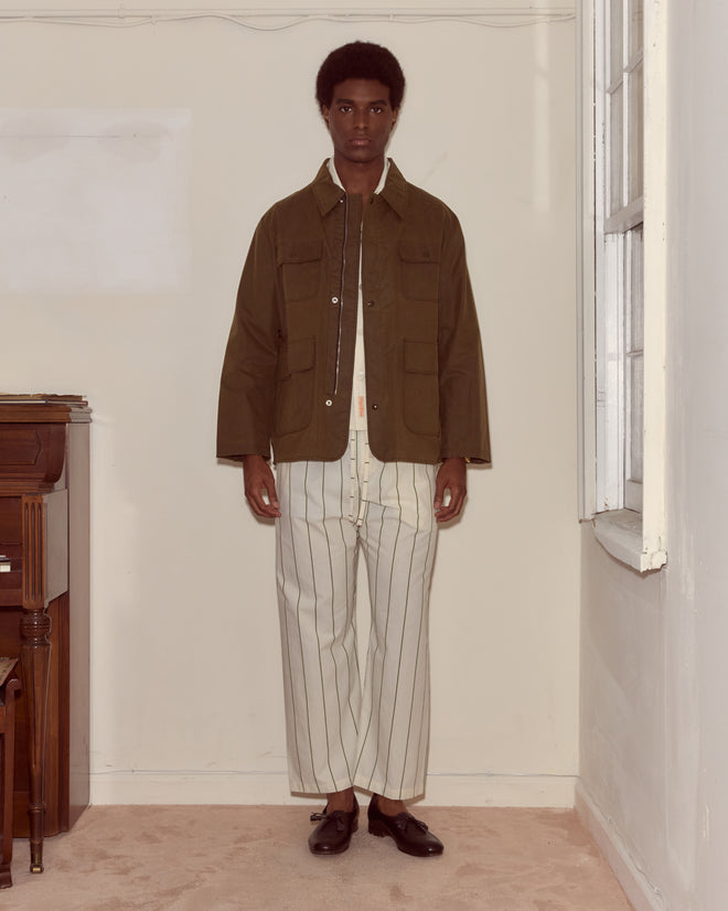 Indoors, a man wears the Waxed Hunting Jacket - Brown, featuring utility pockets, paired with wide cream pants with vertical black stripes.