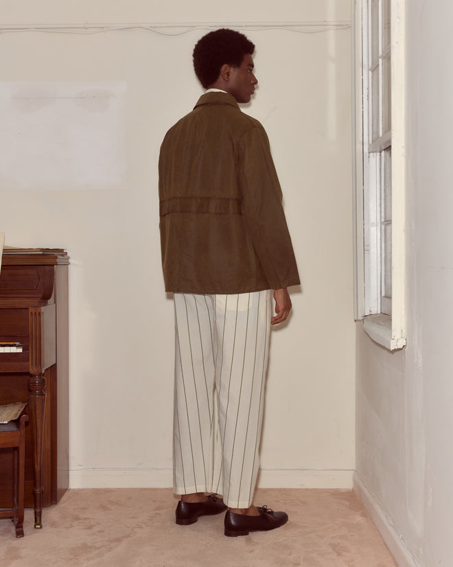 Wearing the Waxed Hunting Jacket - Brown and striped pants, a person stands indoors by a window and piano, facing away.