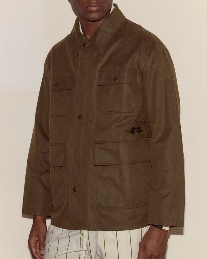 Person wearing a brown Waxed Hunting Jacket with large utility pockets and striped pants, standing against a plain wall.