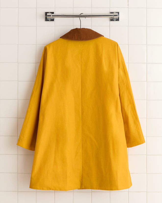 The Waxed Peacoat - Yellow, featuring a waxed cotton finish and brown collar, hangs on a hook against a white tiled wall.
