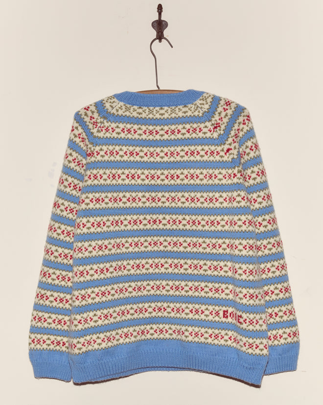 The Wedgewood Sweater, a vintage knitwear piece featuring blue, red, and cream patterns, hangs on a hook against a plain wall.
