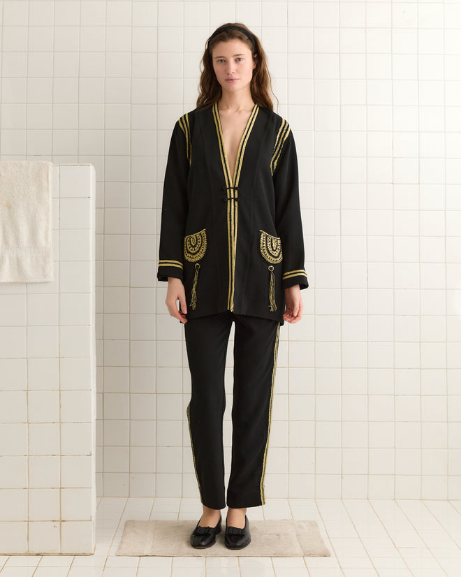 Dressed in a Weedy Crochet Jacket, a person in vintage 1920s-inspired black and gold elegance stands gracefully on a white tile floor, complemented by the white wall's timeless appeal.
