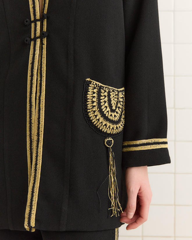 A Weedy Crochet Jacket, a black piece with intricate gold embroidery and tassel details, exudes 1920s-inspired elegance on a standing person.