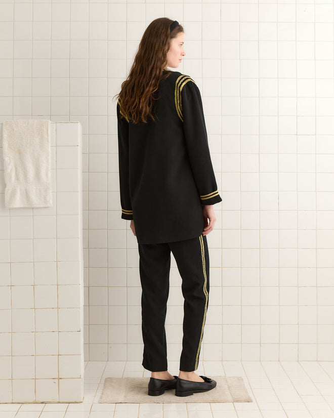 In a white-tiled room, a person stands with their back turned, wearing a black Weedy Crochet Jacket adorned with gold stripes reminiscent of 1920s-inspired fashion. A towel hangs on the wall nearby.