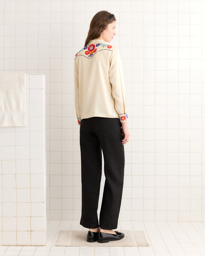 A woman stands on a rug in a tiled room, wearing the Garden Party Western Shirt—a cream top with floral embroidery and pearl snap buttons—paired with black pants.