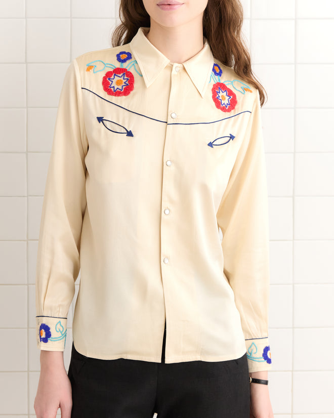 A woman wearing the Garden Party Western Shirt, featuring cream-colored fabric, floral embroidery, and pearl snap buttons, stands against a tiled wall.