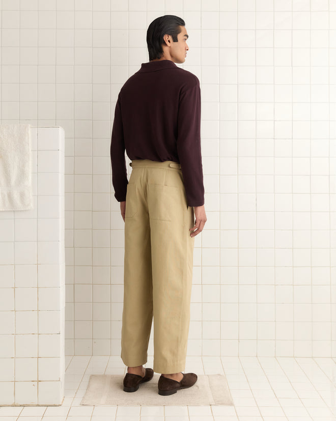 A person with dark hair in a burgundy shirt and khaki wide leg snap trousers stands in a tiled room, facing away.