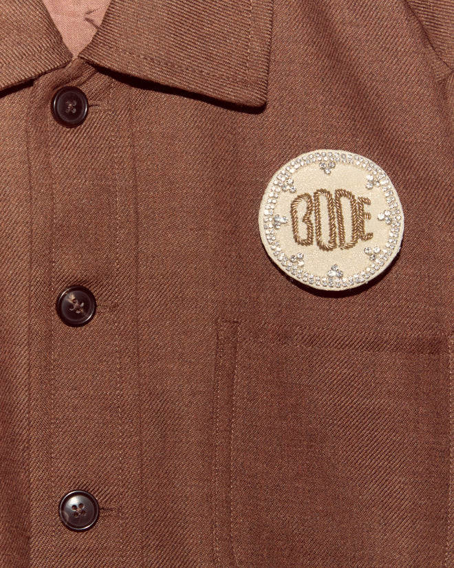 The Willow Brooch - White is a brown shirt with black buttons and a round beige patch reading