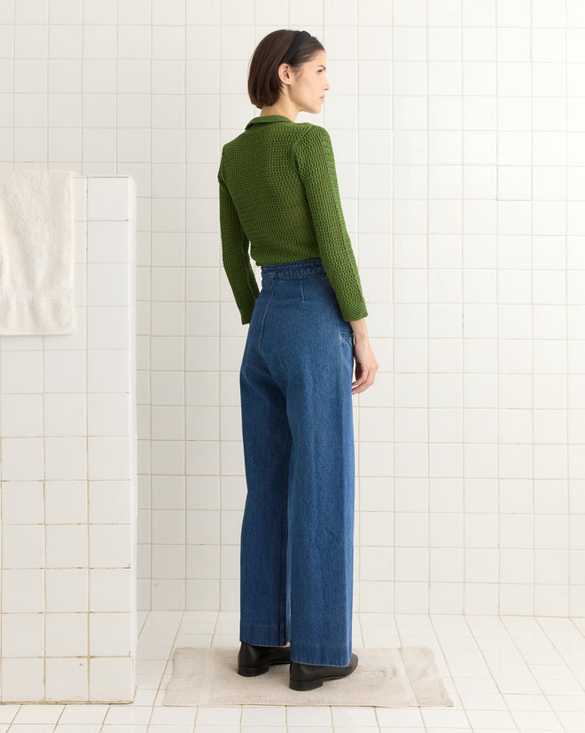 Someone wearing the Willows Pullover in green along with blue jeans stands in a tiled room, viewed from behind.