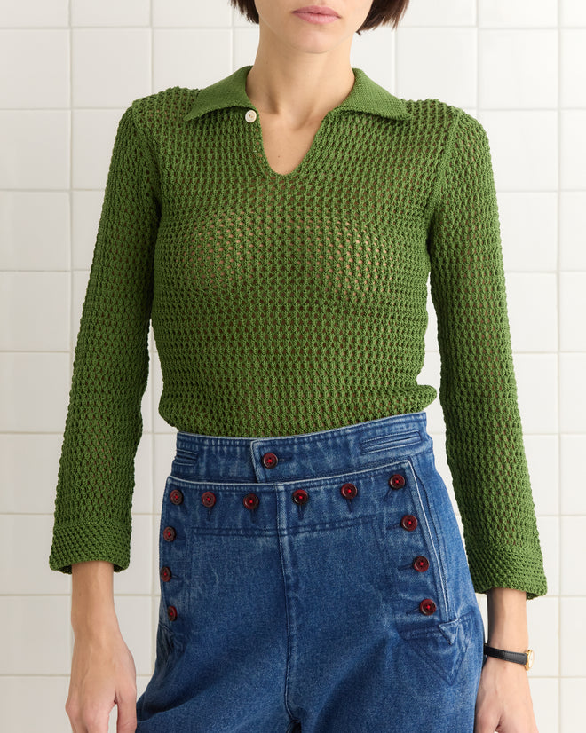 Person wearing the Willows Pullover - Green and high-waisted blue denim pants with red buttons, standing against a tiled wall.