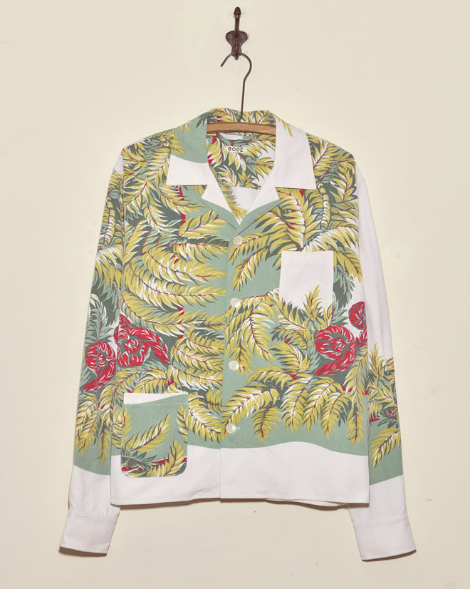 The Windy Fronds Shirt - S/M, featuring unique green, yellow, and red leaf patterns, hangs on a hook against a cream wall.