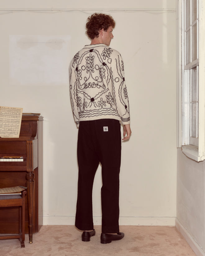 A man stands facing a wall in a room with a piano, wearing the Winter Garden Cardigan and black pants.