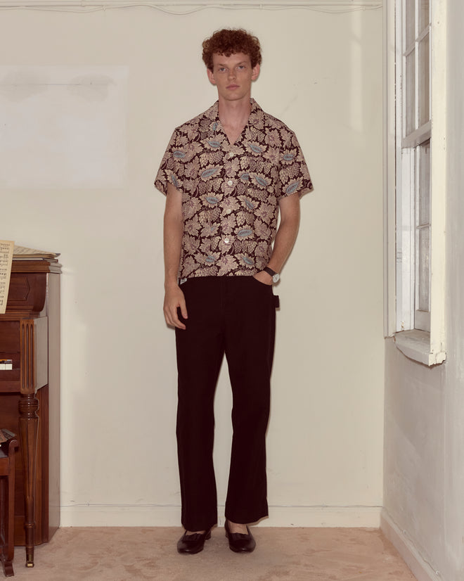 A man with curly hair stands indoors near a piano, wearing the Wisteria Short Sleeve Shirt with a floral print, paired with black pants and black shoes.