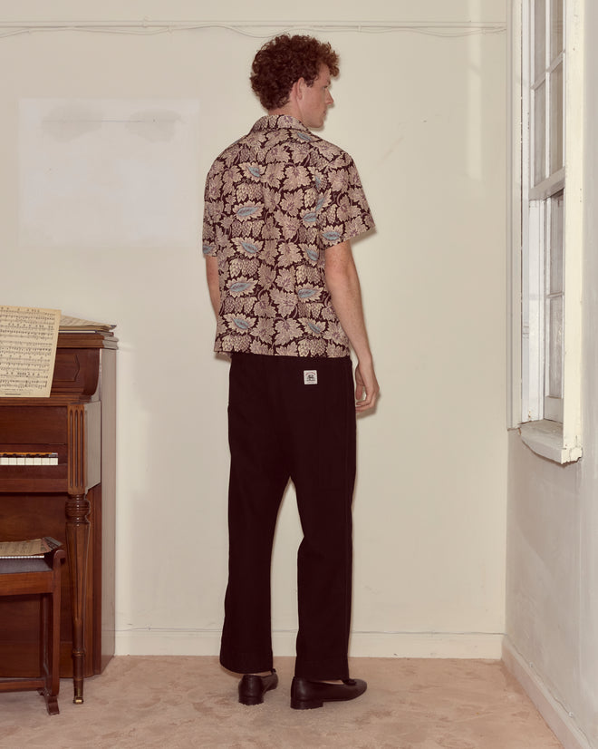 A man in a Wisteria Short Sleeve Shirt and black pants stands facing a corner by a piano and window.