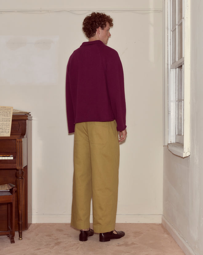 A person wears the Wool Dock Pullover in maroon paired with tan pants, standing by a cream wall near a window and a piano.