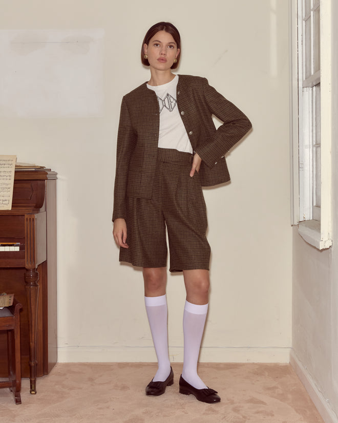 A woman stands indoors by a piano wearing Morris Houndstooth Shorts with D-ring waist adjusters, a plaid blazer, white shirt, knee-high socks, and black shoes.