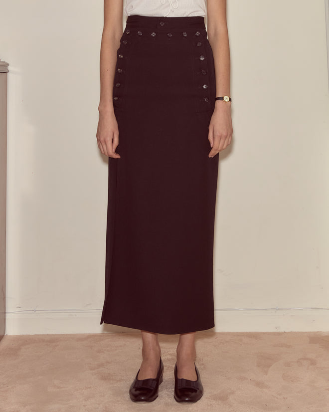 A person wearing a Riding Twill Sailor Skirt, white sleeveless top, black shoes, and a watch stands indoors.