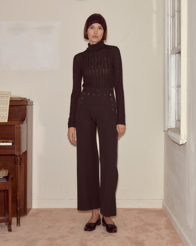 A woman stands indoors wearing a black turtleneck, Riding Twill Sailor Trousers - Black, a black headband, and black shoes.