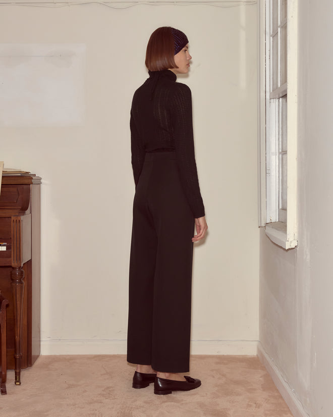 A woman wearing Riding Twill Sailor Trousers in black stands sideways in a sparsely furnished room with beige carpet and white walls.