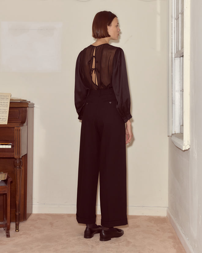 A woman wearing Astor Port Trousers—high rise with wide legs—and a sheer black top with back ties stands facing a wall in a room with a piano.